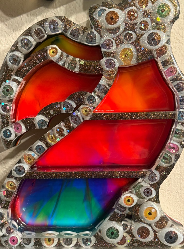 Colorful decorative art piece featuring vibrant red, orange, and green sections, framed with a glittery border and surrounded by eye motifs.